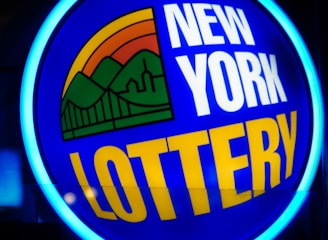 A bright, circular neon sign with a blue background, featuring the words 'New York Lottery' in bold white and yellow letters. The sign includes a colorful illustration of a landscape with green mountains, an orange-red sunset, and a bridge, all encapsulated within the circle.