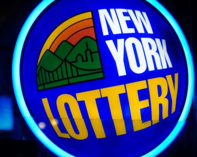 A bright, circular neon sign with a blue background, featuring the words 'New York Lottery' in bold white and yellow letters. The sign includes a colorful illustration of a landscape with green mountains, an orange-red sunset, and a bridge, all encapsulated within the circle.
