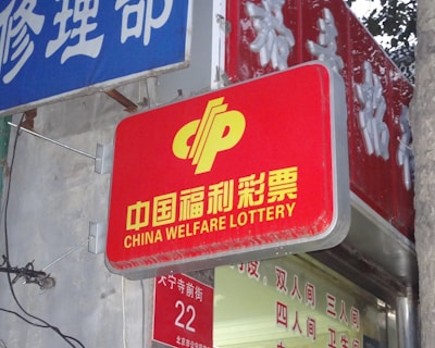 A red sign displaying the text 'China Welfare Lottery' in both Chinese and English, mounted on the exterior of a building. The surroundings include a blue sign with Chinese characters and a partial tree trunk visible on the right side.