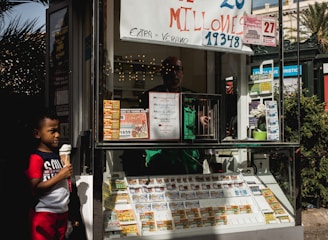A child holding an ice cream cone stands outside a small kiosk or lottery ticket booth. The booth displays various colorful lottery tickets in the window. There are signs advertising a significant lottery prize amount in Spanish. A person can be seen partially obscured inside the booth, and plants are visible around the area, suggesting an outdoor setting.