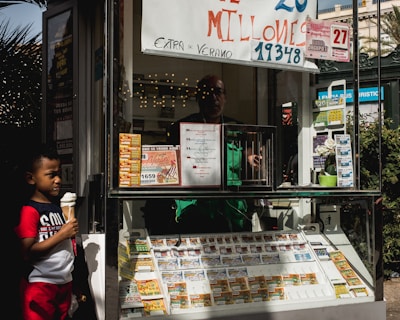 A child holding an ice cream cone stands outside a small kiosk or lottery ticket booth. The booth displays various colorful lottery tickets in the window. There are signs advertising a significant lottery prize amount in Spanish. A person can be seen partially obscured inside the booth, and plants are visible around the area, suggesting an outdoor setting.