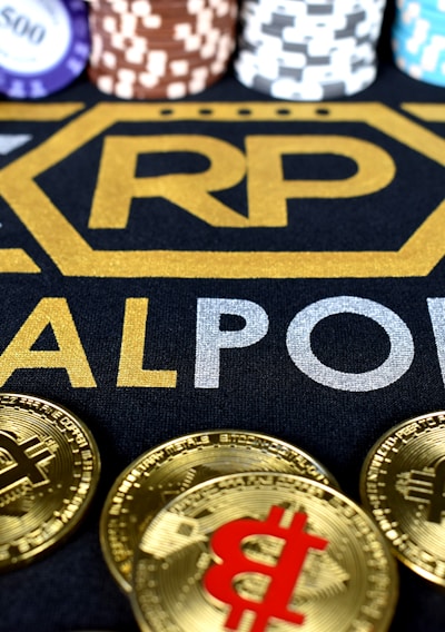 Stacks of poker chips in various colors are arranged in the background, while in the foreground, there are numerous golden coins featuring the Bitcoin symbol. The text 'REALPOKER' is prominently displayed in gold and silver lettering on a dark background.