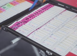 A collection of lottery tickets is organized neatly in a metal tray. The tickets display various numbers and have color-coded sections, primarily in pink and white. Some tickets have vibrant green sections, adding contrast to the overall layout.