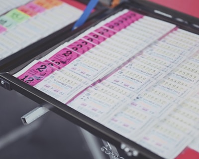 A collection of lottery tickets is organized neatly in a metal tray. The tickets display various numbers and have color-coded sections, primarily in pink and white. Some tickets have vibrant green sections, adding contrast to the overall layout.