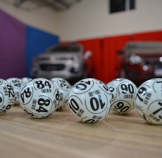 A collection of white lottery balls with black numerals scattered on a wooden surface. Two vehicles are blurred in the background, parked in a room with multicolored walls including teal, purple, blue, and red panels.