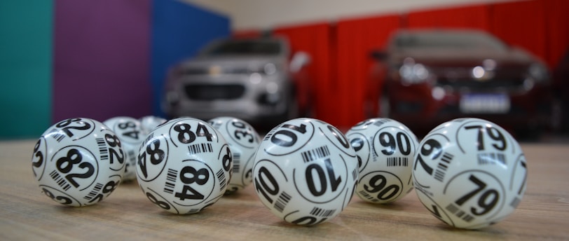 A collection of white lottery balls with black numerals scattered on a wooden surface. Two vehicles are blurred in the background, parked in a room with multicolored walls including teal, purple, blue, and red panels.