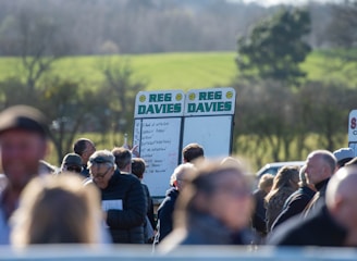 A group of people gathered outdoors with two large betting boards in the background labeled 'REG DAVIES'. The setting appears to be a countryside location with trees and fields visible in the distance. The crowd consists of individuals dressed in jackets and hats, engaging in conversation.