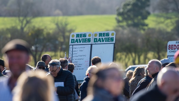 A group of people gathered outdoors with two large betting boards in the background labeled 'REG DAVIES'. The setting appears to be a countryside location with trees and fields visible in the distance. The crowd consists of individuals dressed in jackets and hats, engaging in conversation.