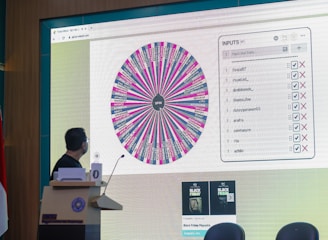A person stands in front of a large screen displaying a colorful digital wheel with names. The screen shows a web interface with options for inputs, featuring a list of names with checkboxes on one side and a large, vibrant spinning wheel on the other. The environment appears to be indoors, possibly in a conference or presentation setting with a podium.